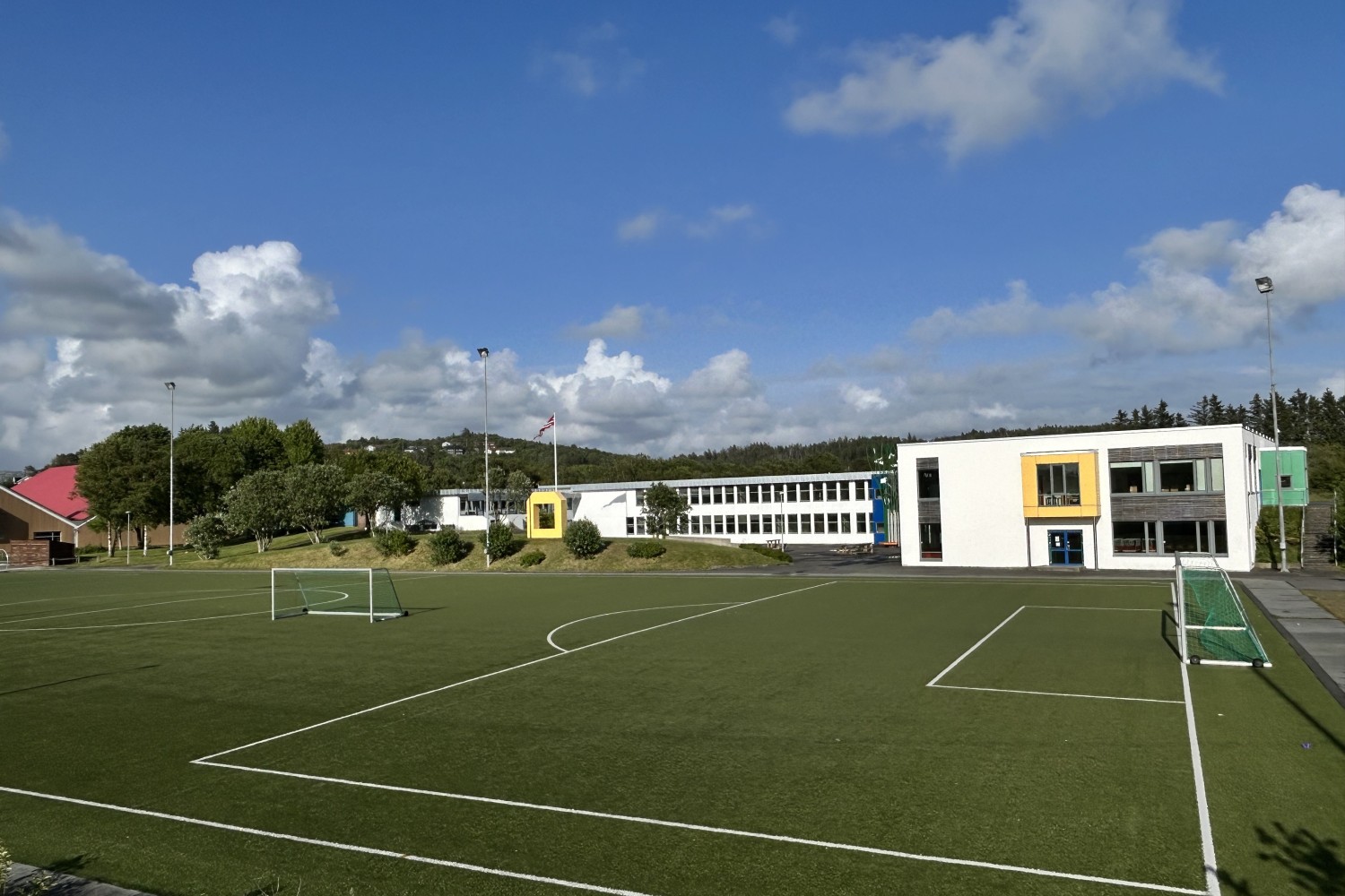 Et stort, grønt kunstgressfotballbane med små mål står i forgrunnen. I bakgrunnen ligger en skole med hvite bygninger, noen med fargede rammer rundt vinduene i gult og grønt. Det er trær og busker rundt området, og et flagg vaier ved en flaggstang. Himmlen er blå med spredte skyer.