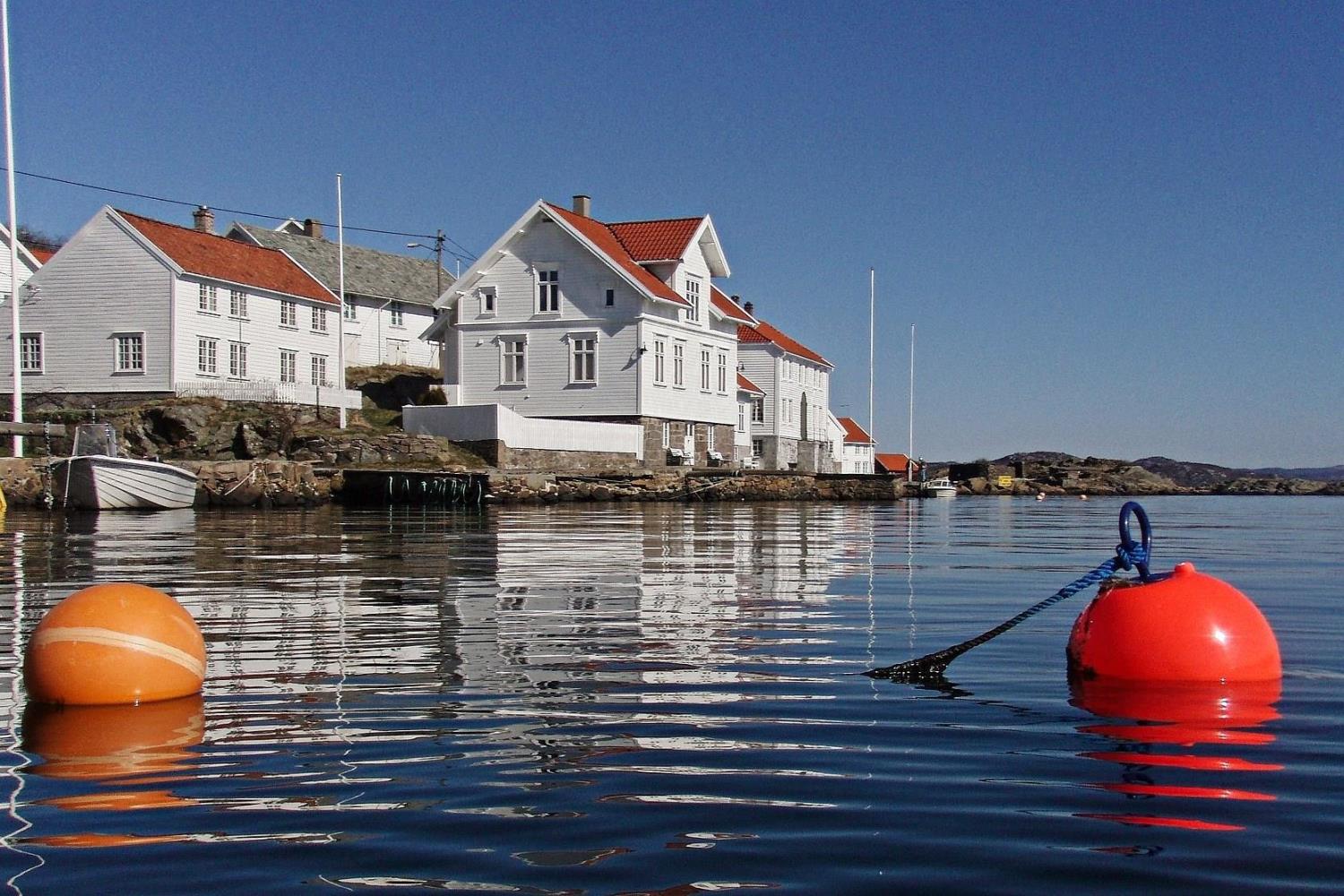 Røde og oransje bøyer flyter i rolig sjø foran hvite trehus med røde tak langs strandkanten i Loshavn, under blå himmel.
