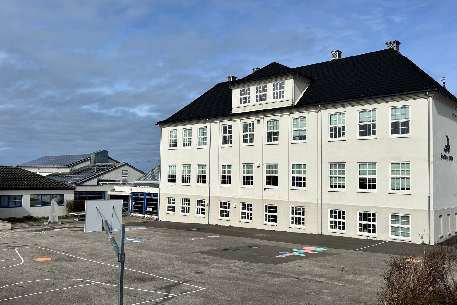 Forsiden av Borhaug skole med en stor, lys skolebygning i tre etasjer. Foran bygningen er en asfaltert skolegård med oppmerkede lekeområder og en basketballkurv. Mindre skolebygg sees til venstre, og himmelen er delvis skyet.
