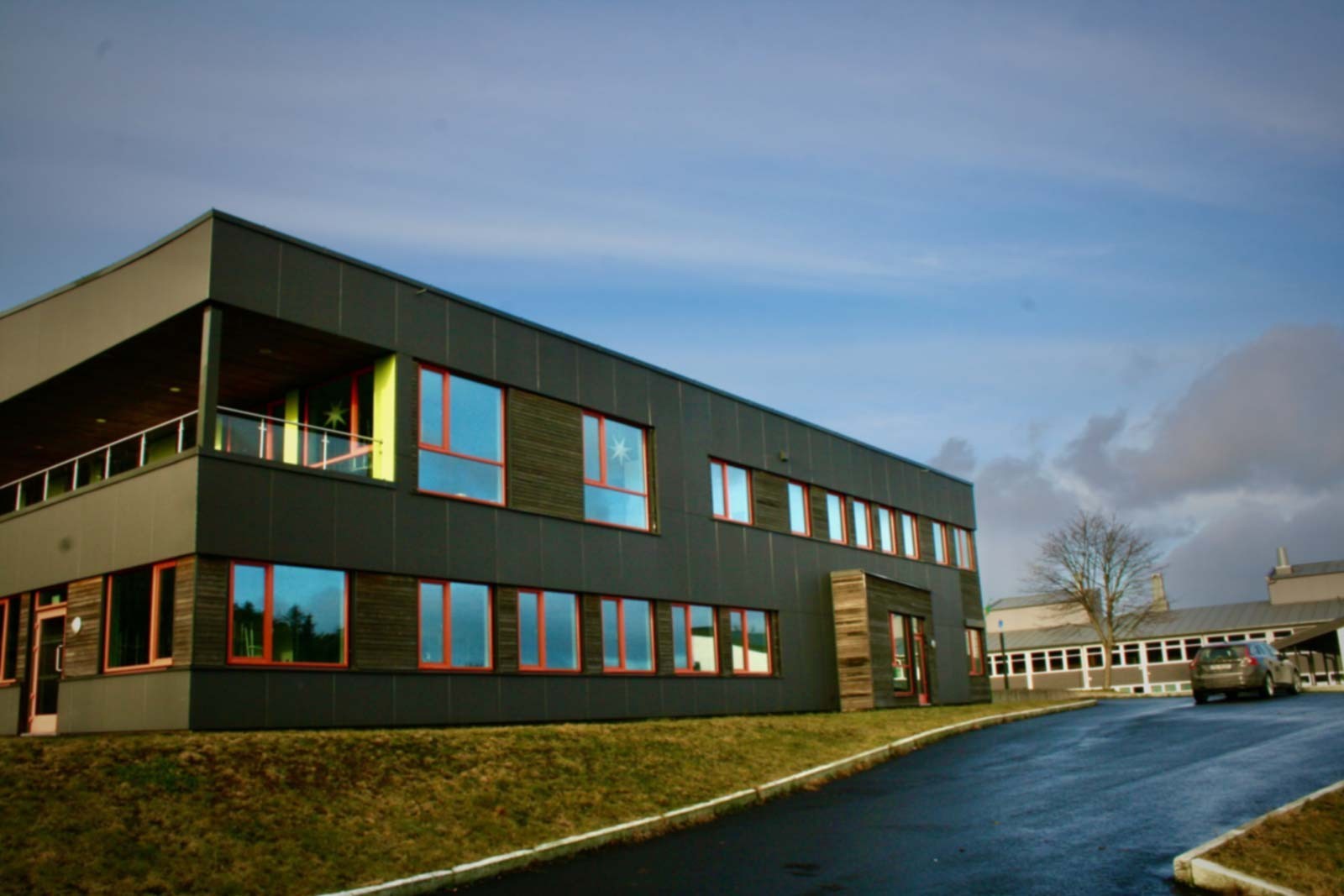 Moderne skolebygning med to etasjer, store vinduer og mørk fasade. Bygget står på en gressbakke ved en asfaltert innkjørsel, med klar himmel og noen skyer i bakgrunnen